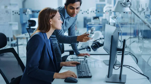 Modernes Fabrikbüro: Männlicher Projektleiter spricht mit einer Industrieingenieurin, die am Computer arbeitet. Professionelle Teamarbeit, junge Spezialisten, die Probleme lösen und den technologischen Fortschritt vorantreiben — Stockfoto