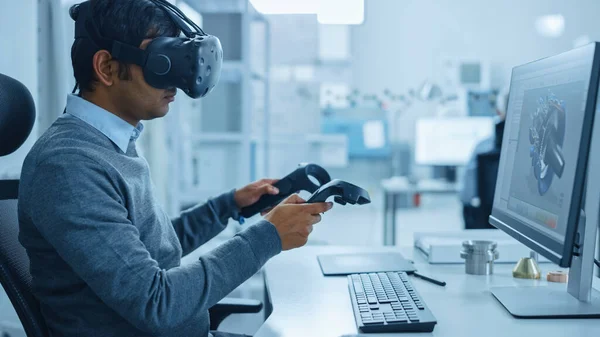 Modern Industrial Factory: Mechanical Engineer Wearing Virtual Reality Headset, Holding Controllers, Uses VR technology for Industrial Design, Development and Prototyping in CAD Software on Computer.