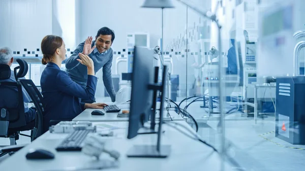 Inside Factory Office: Man Project Supervisor samtalar med en kvinnlig industriell ingenjör som arbetar på dator, ge hög fem. I verkstad: Arbetstagare använder högteknologisk industri 4 CNC-maskiner, Robot Arm. — Stockfoto