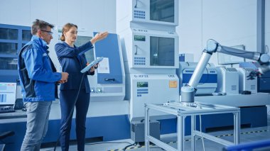 Modern Factory: Kadın Proje Yöneticisi ve Erkek Mühendis Ayakta, Konuşan ve Tablet Bilgisayar kullanarak Robot Kolu Toplama Hattı Programlama. High Tech CNC ile Çağdaş Tesis. İndsutry 4.0