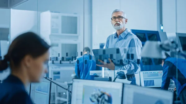 Fábrica moderna: Ingeniero sénior mira la cámara, sostiene el ordenador portátil, inspecciona, supervisa la operación del taller. Manufactory tiene profesionales trabajando en maquinaria CNC, brazo de robot . — Foto de Stock