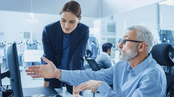 Bureau d'usine moderne Une jeune gestionnaire de projet confiante s'entretient avec un ingénieur principal travaillant sur ordinateur. Équipe de professionnels Résoudre les problèmes de l'industrie lourde, avoir des discussions, pointer du doigt — Photo