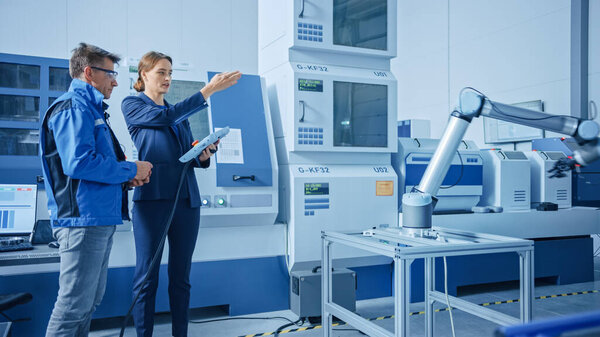 Modern Factory: Female Project Manager and Male Engineer Standing, Talking and Using Tablet Computer for Programming Robot Arm Assembly Line. Contemporary Facility with High Tech CNC. Indsutry 4.0