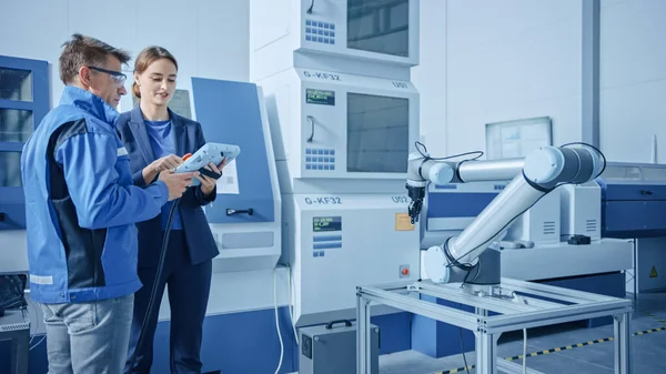 Fábrica moderna: Gerente de projeto feminino e Engenheiro Masculino em pé, conversando e usando o Tablet Computer para programar a linha de montagem do braço robô. Instalação Contemporânea com Máquinas CNC de Alta Tecnologia — Fotografia de Stock