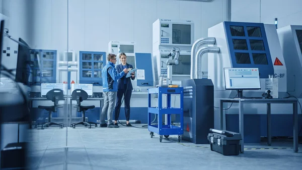Modern Factory: Kadın Proje Yöneticisi ve Erkek Mühendis Ayakta, Konuşan ve Tablet Bilgisayar kullanarak Robot Kolu Toplama Hattı Programlama. Yüksek Teknoloji CNC Makineleri ile Çağdaş Tesis