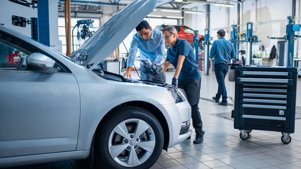 Manager controleert diagnostiek Resultaten op een tablet computer en verklaart een uitsplitsing van de motor naar een vrouwelijke monteur. Auto Service Medewerkers Inspecteren Cars Engine Bay met een LED-lamp. Moderne Clean Workshop. — Stockfoto