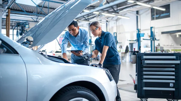 Manager comprueba los resultados de diagnóstico en un Tablet Computer y explica un fallo del motor a una mecánica femenina. Empleados de servicio de automóviles inspeccionan la bahía del motor de coches con una lámpara led. Taller limpio moderno . — Foto de Stock