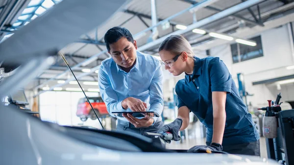 Instructeur avec un ordinateur tablette donne une tâche pour un futur mécanicien. Une étudiante inspecte le moteur de voiture. Assistant vérifie la cause d'une panne dans le véhicule dans un service de voiture. — Photo