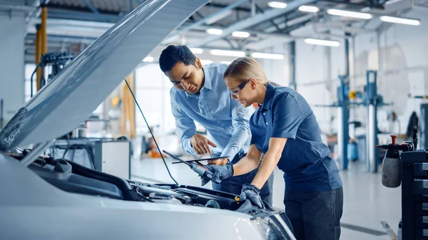 Instructor con un Tablet Computer está dando una tarea para un futuro mecánico. Estudiante femenina inspecciona el motor del coche. Asistente está comprobando la causa de una avería en el vehículo en un servicio de coche . — Foto de Stock