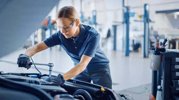Hermoso empoderamiento mecánico femenino está trabajando en un coche en un servicio de coche. Mujer con gafas de seguridad está trabajando en un mantenimiento habitual del coche. Usa un trinquete. Taller limpio moderno con coches . —  Fotos de Stock