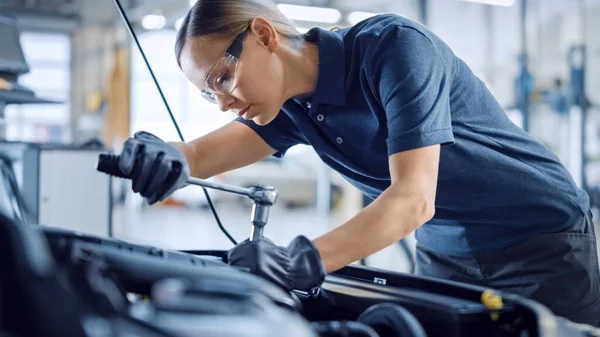 Vacker Empowering kvinnliga mekaniker arbetar på en bil i en bil service. Kvinnan i säkerhetsglas fixar motorn. Hon använder en Ratchet. Modern Clean Workshop med bilar. — Stockfoto