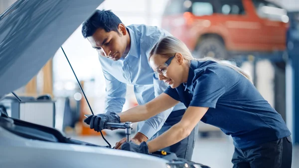 Le gestionnaire vérifie les résultats du diagnostic sur un ordinateur tablette et explique une panne moteur à une mécanicienne. Employé de service de voiture utilise un cliquet pour fixer la composante moteur. Atelier moderne propre. — Photo