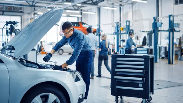 Mecánico profesional está trabajando en un coche en un servicio de coche. Reparador en gafas de seguridad está trabajando en un mantenimiento habitual del coche. Él cuelga una lámpara y trabaja con un trinquete. Taller limpio moderno . — Foto de Stock