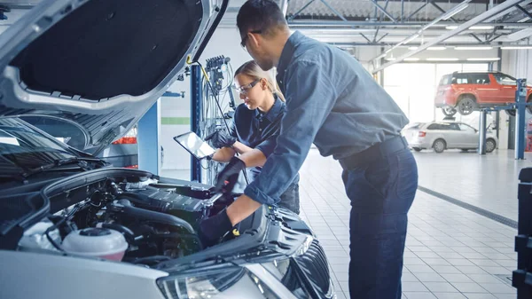 Dois mecânicos em um serviço estão inspecionando um carro depois que obtiveram os resultados do diagnóstico. Especialista feminina está comparando os dados em um computador tablet. Reparador está usando um catraca para reparar as falhas . — Fotografia de Stock