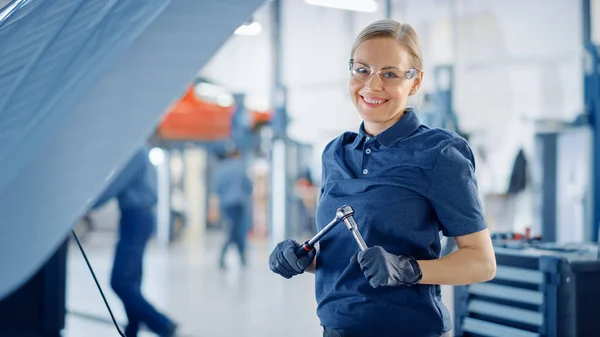 Piękne upoważnienie Kobieta mechanik samochodowy pracuje nad pojazdem w serwisie. Wygląda na szczęśliwą, gdy używa grzechotki. Specjalista nosi okulary ochronne. Patrzy w kamerę i uśmiecha się.. — Zdjęcie stockowe