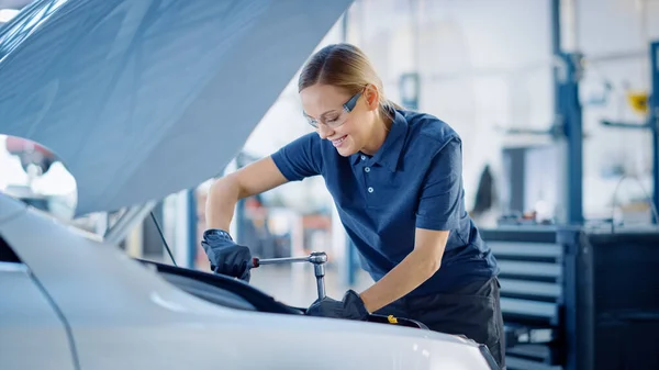 Mooie empowerment vrouwelijke auto monteur werkt aan een voertuig in een dienst. Ze ziet er gelukkig uit terwijl ze een ratel gebruikt. Specialist is het dragen van veiligheidsbril. — Stockfoto