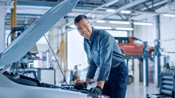 Handsome mecánico profesional del coche está trabajando en un vehículo en un servicio. El reparador se ve feliz mientras usa un trinquete. Especialista está usando gafas de seguridad. Mira una cámara y sonríe . — Foto de Stock
