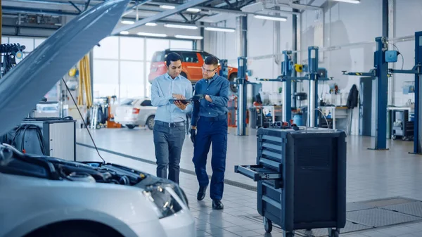 Le gestionnaire vérifie les résultats du diagnostic sur un ordinateur tablette et explique une panne de véhicule à un beau mécanicien. Les employés du service d'auto parlent en marchant dans un garage. Atelier moderne propre. — Photo