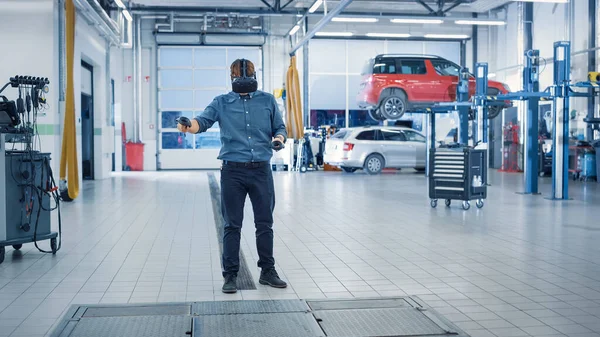 Especialista está vestindo um fone de ouvido de realidade virtual e segurando dois Joysticks em um serviço de carro. Mecânico está investigando a ruptura com a ajuda da tecnologia futura. Reparador em oficina moderna . — Fotografia de Stock