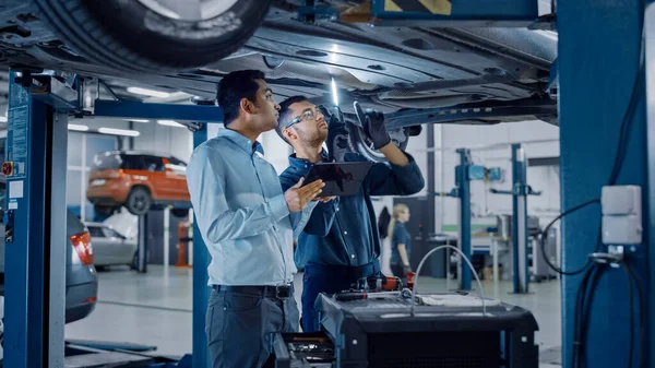 Manager überprüft Daten auf einem Tablet-Computer und erklärt die Aufschlüsselung zu einem Mechaniker. Mitarbeiter des Autoservice untersuchen den Boden des Autos mit einer LED-Lampe. Moderne saubere Werkstatt. — Stockfoto