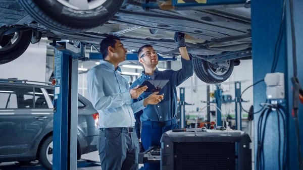 Manager comprueba los datos en un Tablet Computer y explica el desglose a un mecánico. Los empleados de servicio de automóviles inspeccionan la parte inferior y las placas de deslizamiento del automóvil. Taller limpio moderno . —  Fotos de Stock