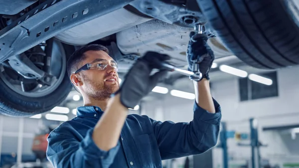 Retrato de un mecánico guapo trabajando en un vehículo en un servicio de coche. El reparador profesional está usando guantes y usando un trinquete debajo del coche. Taller limpio moderno. — Foto de Stock