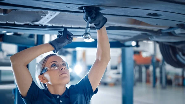 Porträtt Skott av en kvinnlig mekaniker som arbetar på ett fordon i en bilservice. Bemyndigande kvinna bära handskar och använda en Ratchet under bilen. Modern Clean Workshop. — Stockfoto