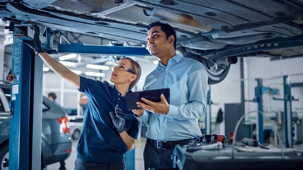 Feminino Mecânico Falando com um gerente sob um veículo em um serviço de carro. Especialista está mostrando informações em um computador tablet. Capacitando mulher vestindo luvas e luvas de segurança. Oficina limpa moderna . — Fotografia de Stock