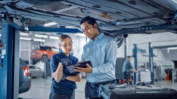 Žena Mechanik mluví s manažerem pod vozidlem v autoservisu. Specialista je zobrazování informací v tabletu počítače. Posílení ženských rukavic a bezpečnostních rukavic. Moderní čistý workshop. — Stock fotografie