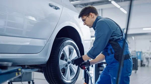 Mecânico em macacão azul é desaparafusar porcas de Lug com uma chave de impacto pneumático. Reparador funciona em um serviço de carro limpo moderno. Especialistas removem a roda para consertar um componente em um veículo . — Fotografia de Stock