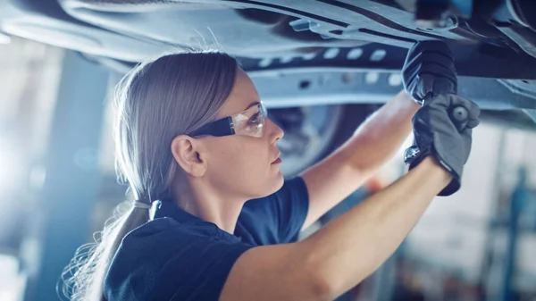 Vackra Porträtt av en professionell kvinnlig bil mekaniker arbetar under ett fordon på en hiss i tjänsten. Hon använder en Ratchet. Specialisten är att bära skyddsglasögon. Modern Clean Workshop. — Stockfoto