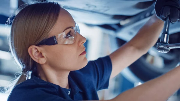 Bellissimo ritratto di un meccanico di auto femminile professionista sta lavorando sotto un veicolo su un ascensore in servizio. Sta usando un cricchetto. Lo specialista indossa occhiali di sicurezza. Laboratorio pulito moderno. — Foto Stock