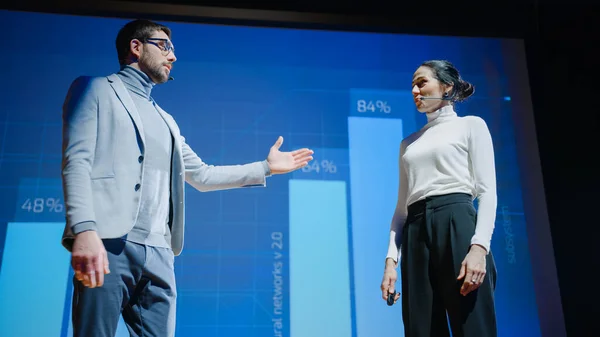 En el escenario, la mujer ejecutiva exitosa y el gerente masculino presentan un nuevo producto, muestran infografías, estadísticas en pantalla grande, hablan sobre el crecimiento. Evento en vivo, Tech Startup, Conferencia de Negocios —  Fotos de Stock