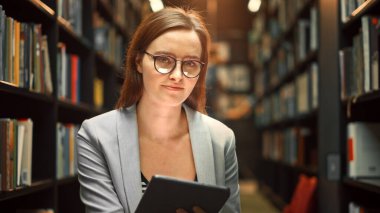 Üniversite Kütüphanesi Çalışması: Kameraya Bakan Güzel Beyaz Kız Gülümseyen Portresi. Yerde oturan tatlı otantik kız, Dijital Tablet Bilgisayarında atama kağıdı yazıyor.