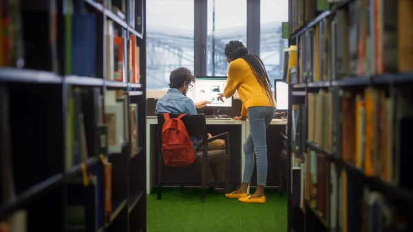 Universitätsbibliothek: Junge benutzt Personal Computer an seinem Schreibtisch, spricht mit einer Klassenkameradin, die ihm bei Klassenaufgaben hilft. Fokussierte Studenten lernen gemeinsam. Schuss zwischen Bücherregalreihen — Stockfoto