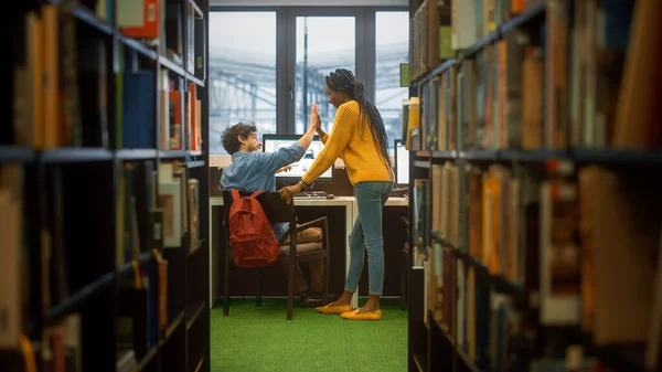 Biblioteca de la Universidad: Niño utiliza la computadora personal en su escritorio, Charlas con compañera de clase Dar Choque Cinco después de Solución exitosa. Estudiantes enfocados estudian juntos. Disparo entre filas de estanterías — Foto de Stock