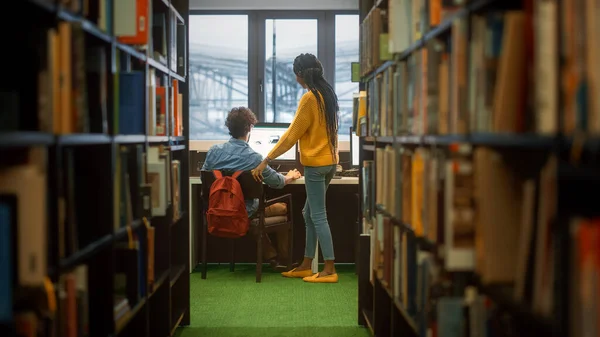 Egyetemi Könyvtár: A fiú személyi számítógépet használ az íróasztalánál, beszél a lány osztálytársával, aki elmagyarázza, segít neki az osztálybeosztásban. Összpontosított diákok együtt tanulnak. Lövöldözés a könyvespolcok sorai között — Stock Fotó