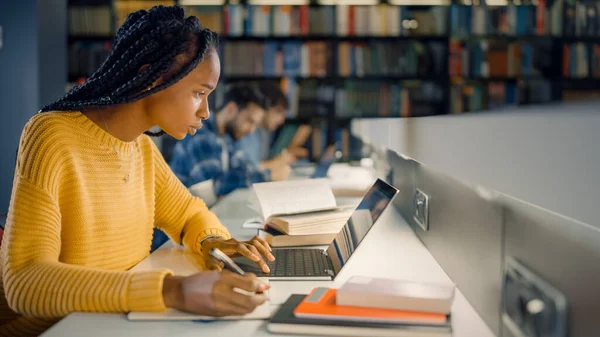 Biblioteca de la Universidad: Chica negra dotada utiliza el ordenador portátil, escribe notas para el papel, ensayo, estudio para la asignación de clases. Diversos grupos multiétnicos de estudiantes que aprenden, estudian para exámenes, hablan en la universidad — Foto de Stock