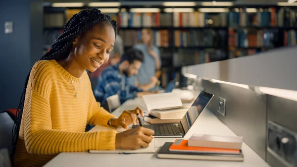 Bibliothèque universitaire : Une fille noire douée utilise un ordinateur portable, écrit des notes pour le papier, un essai, une étude pour une affectation en classe. Divers groupes multi-ethniques d'étudiants Apprentissage, Étudier pour les examens, Parler au collège — Photo