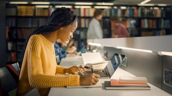 Biblioteca de la Universidad: Hermosa chica negra dotada sentada en el escritorio, utiliza el ordenador portátil, escribe notas para el papel, ensayo, estudio para la asignación de clases. Diverso grupo de estudiantes aprendiendo, estudiando para exámenes . — Foto de Stock