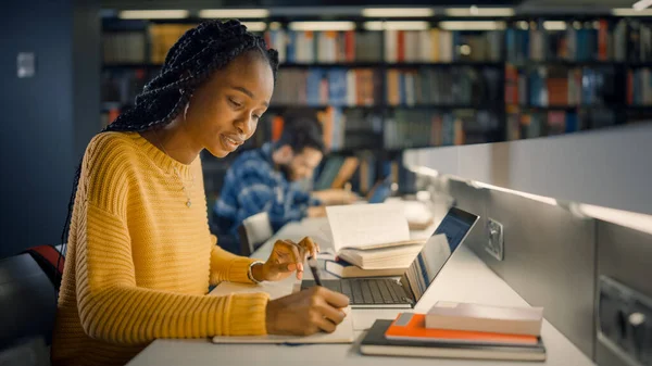 Biblioteca de la Universidad: Hermosa chica negra dotada sentada en el escritorio, utiliza el ordenador portátil, escribe notas para el papel, ensayo, estudio para la asignación de clases. Diverso grupo de estudiantes aprendiendo, estudiando para exámenes . —  Fotos de Stock