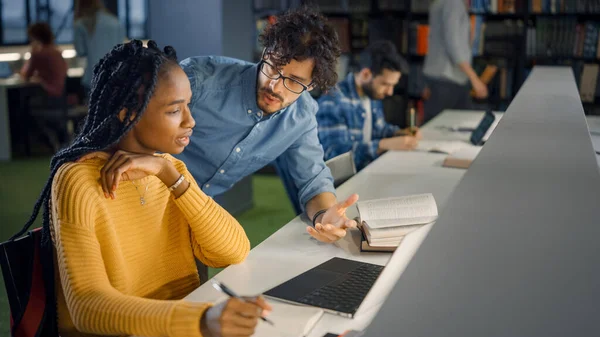 Bibliothèque universitaire : Une fille noire douée utilise un ordinateur portable, un camarade de classe intelligent l'explique et l'aide dans son devoir de classe. Heureux étudiants diversifiés Parler, apprendre, étudier ensemble pour les examens — Photo