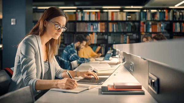 Biblioteca de la Universidad: Talentosa niña caucásica sentada en el escritorio, utiliza el ordenador portátil, escribe notas para el papel, ensayo, estudio para la asignación de clases. Diverso grupo de estudiantes aprendiendo, estudiando para exámenes . —  Fotos de Stock