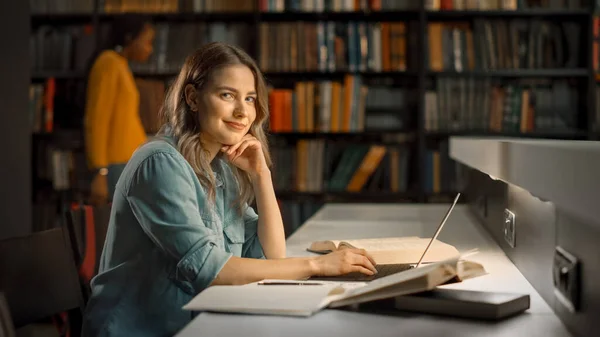 Biblioteca Universitária: A menina branca brilhante focada trabalha na atribuição, usa o computador, olha para a câmera e sorri. Menina inteligente bonito com individualidade outnehtic — Fotografia de Stock