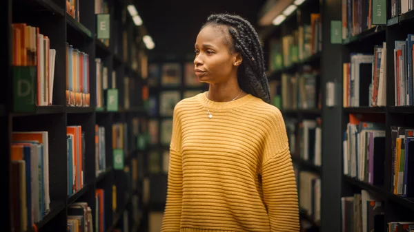 大学图书馆：在各排书架之间寻找合适的课题名称的精美黑人女生肖像 — 图库照片