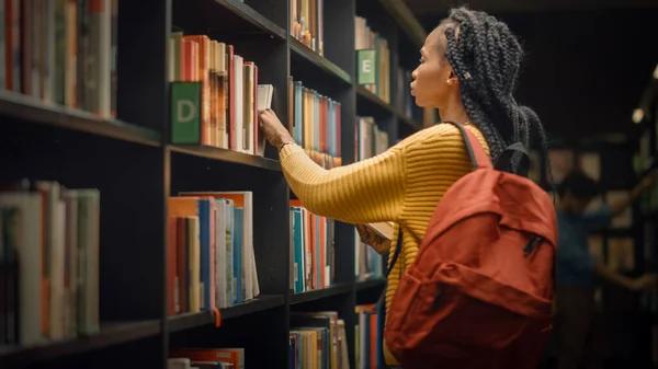 Університетська бібліотека: Портрет обдарованої красивої чорної дівчини стоїть між рядками книжкових полиць, використовуючи смартфон, шукаючи правильну назву книги, знаходить і вибирає одну для призначення класу — стокове фото