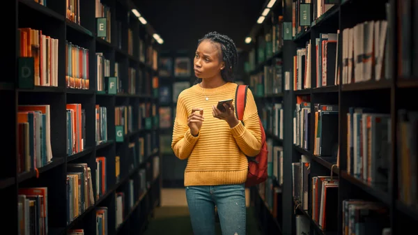 University Library: Portrait of Gifted Beautiful Black Girl Stands Between Rows of Bookshelves Using Smartphone Searching for the Right Book Title for Class Assignment