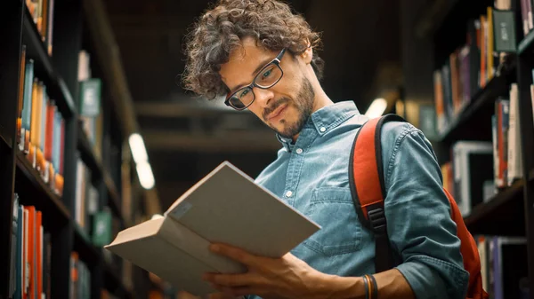 Universiteitsbibliotheek: Talentvolle Latijns-Amerikaanse jongen dragen van glazen staande naast boekenplank Leest boek voor zijn klasse opdracht en examenvoorbereidingen. Lage hoek Portret — Stockfoto
