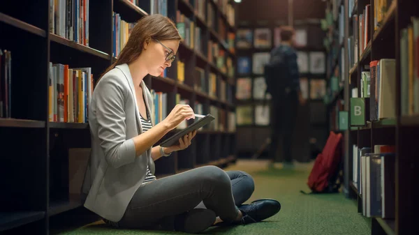 대학 도서관: 아름다운 캅카스 소녀 가 바닥에 앉아 있는 모습, 사용하는 디지털 태블릿 컴퓨터 , Writes Notes, Study for Class Assignment. 다양 한 학생들 이 공부하고 있으며, 연구하고 있다 — 스톡 사진