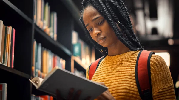 Biblioteca universitaria: Smart Bella ragazza nera in piedi accanto a Libreria Holding e lettura del libro di testo, facendo ricerca per il suo compito di classe e preparativi esame. Ritratto ad angolo basso — Foto Stock